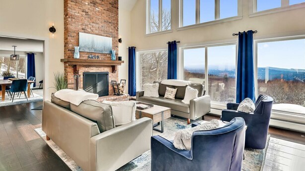 A spacious living room with a brick fireplace, large windows, and a view of the outdoors. - Stoweic Views  - Luxury Dream Vacation Home (Stowe)
