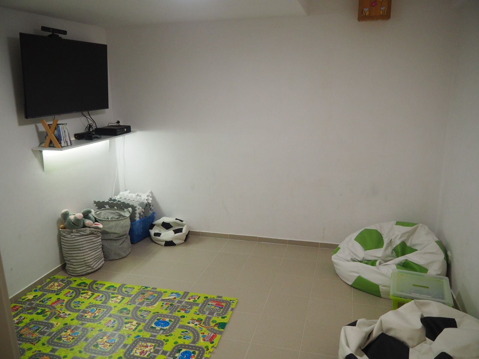 A small room with a TV mounted on the wall, a shelf with books and a small lamp, a colorful mat on the floor, and a bean bag chair.