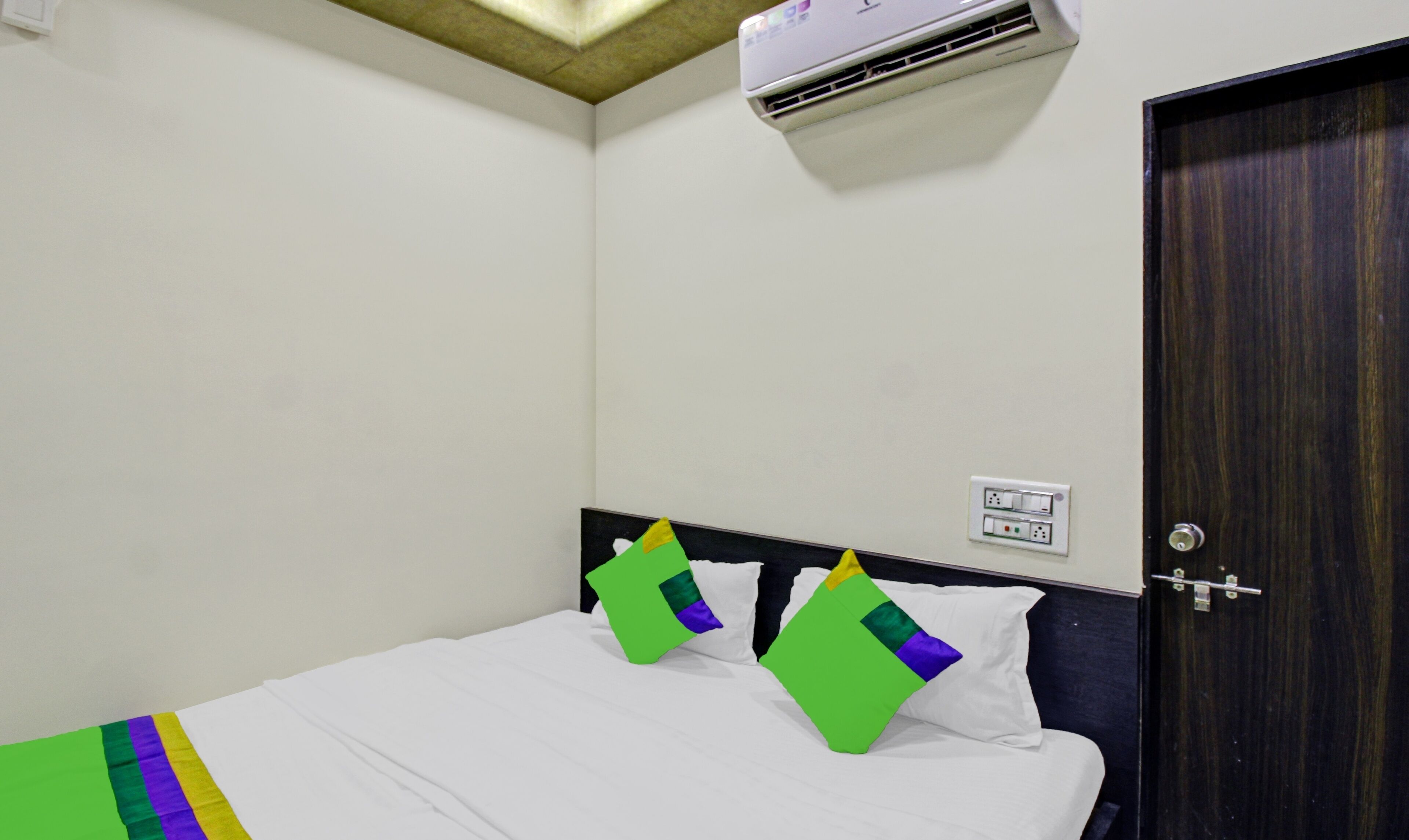 A hotel room with a bed, a wall-mounted air conditioner, a door, and a power outlet.