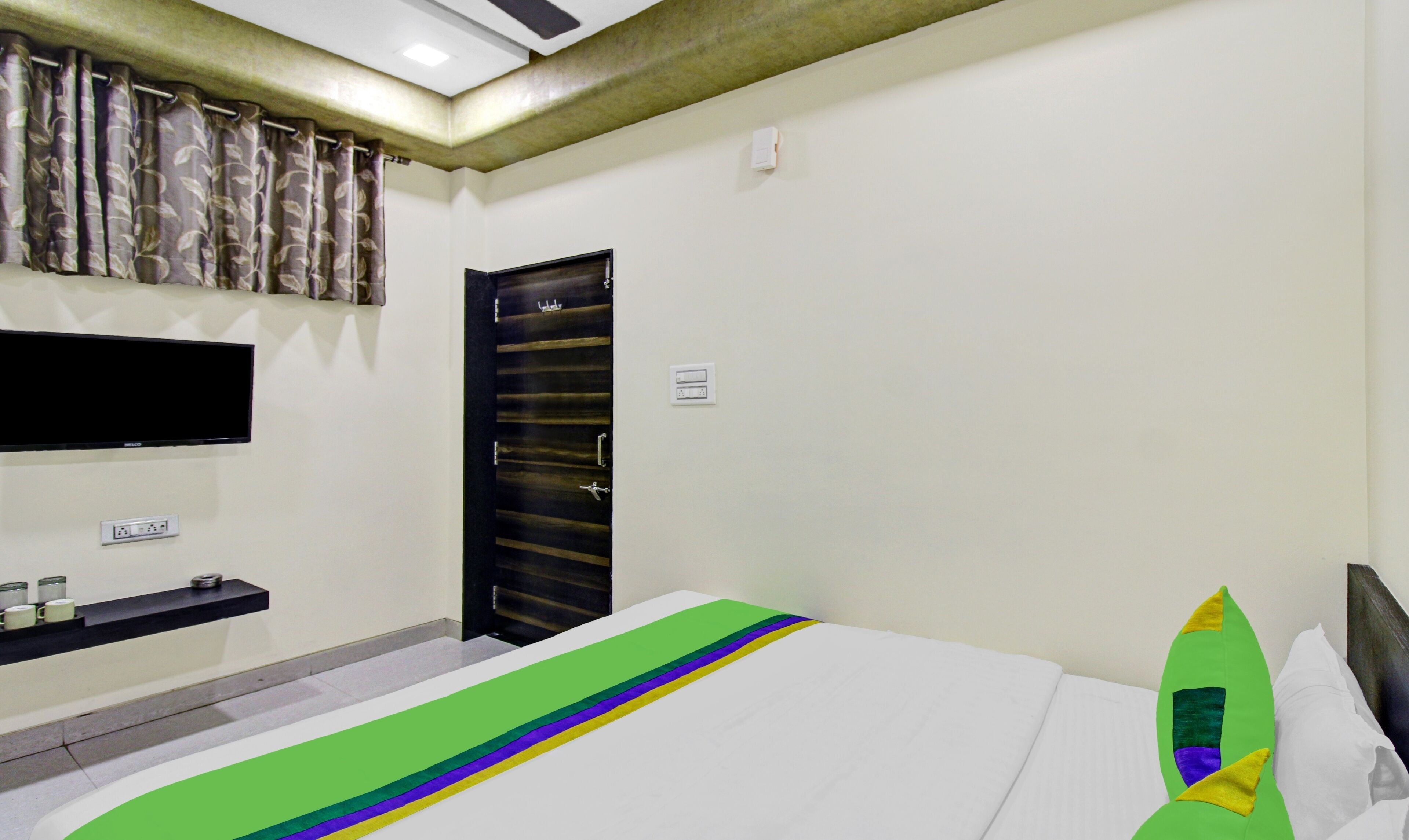 A hotel room with a bed, a TV mounted on the wall, a bathroom with a shower curtain, and a door.