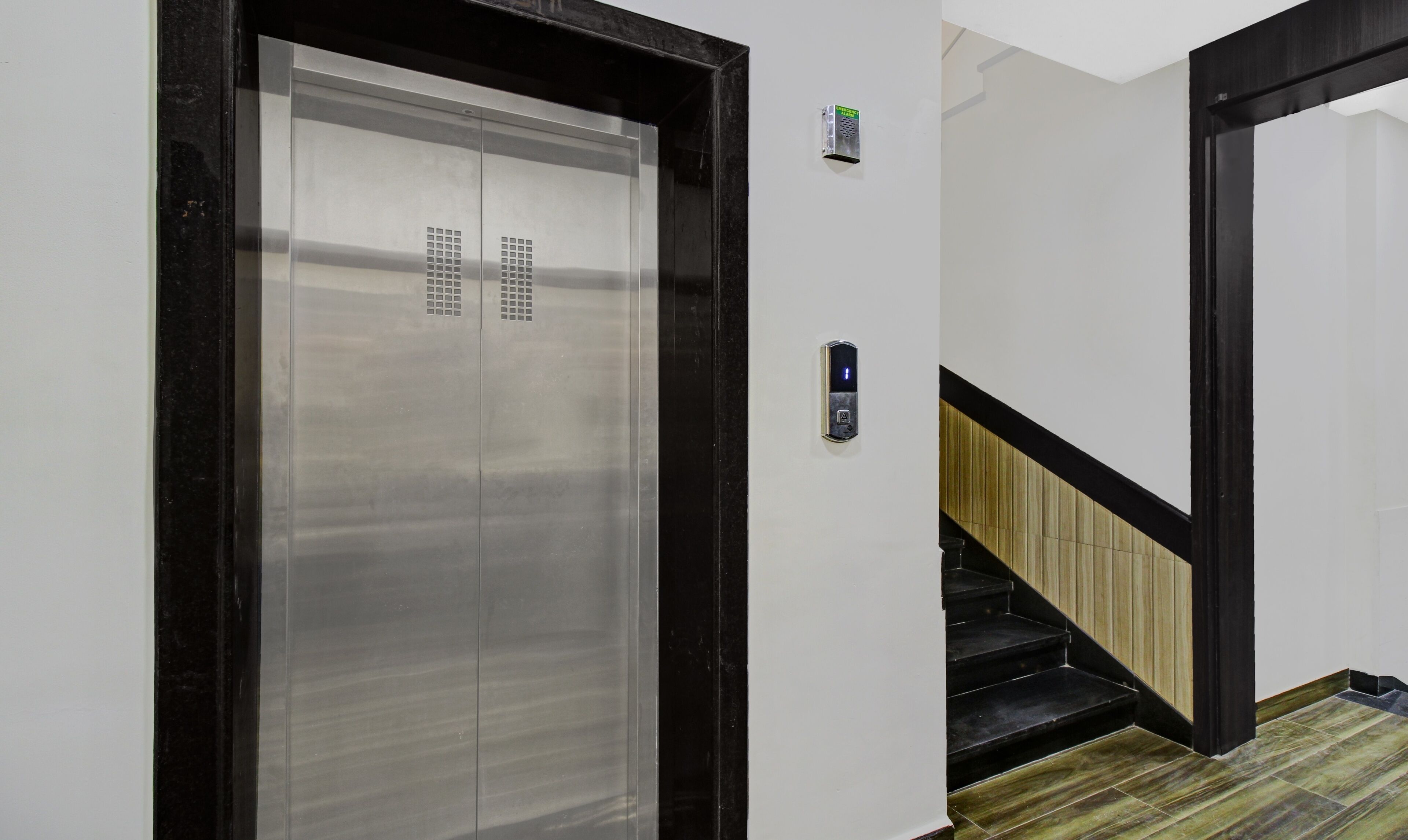 An elevator with a metallic finish and a black frame, a wall-mounted intercom, and a staircase with wooden steps and black railings.