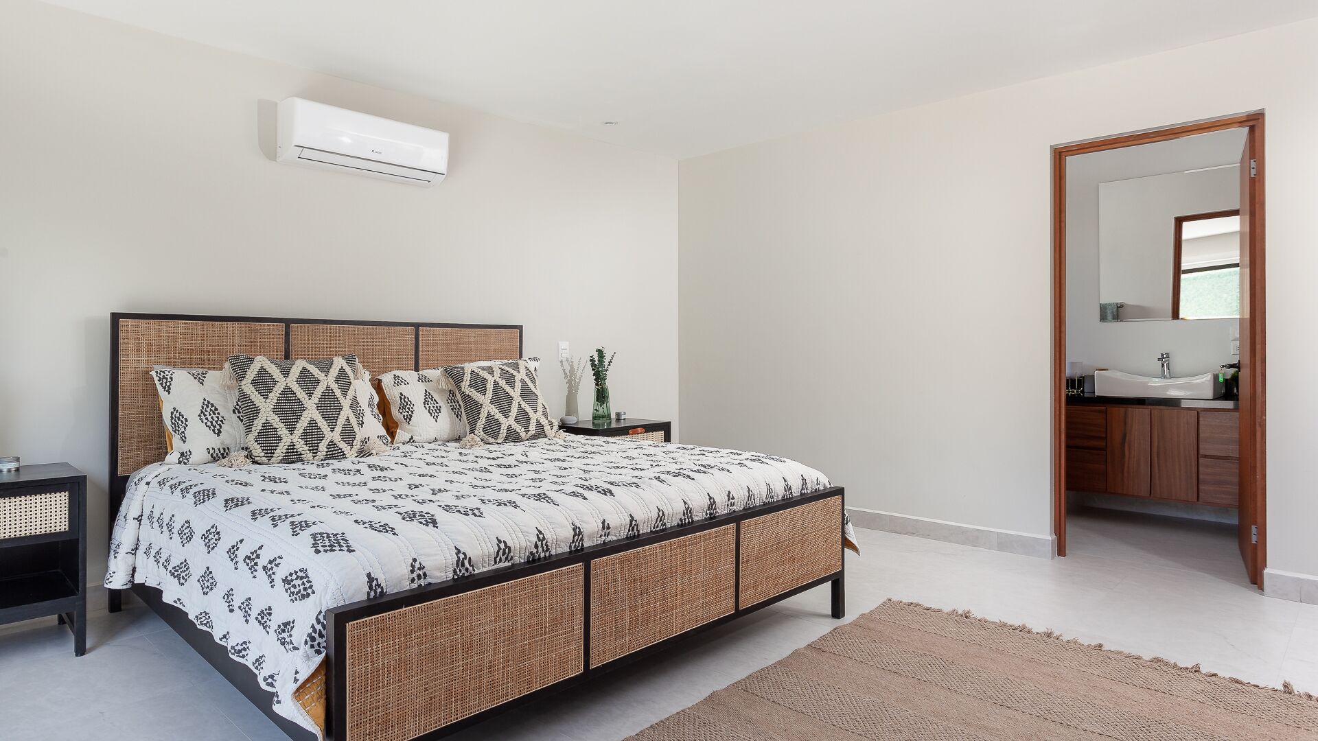 A bedroom with a bed, bedside tables, a bathroom visible through an open door, and an air conditioning unit on the wall.