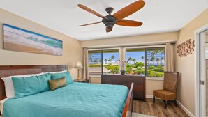 A bedroom with a bed, a ceiling fan, a desk with a chair, a window with a view of palm trees, and a painting of a beach. - Tropical Oasis - Stunning Views In Private Setting (Princeville)