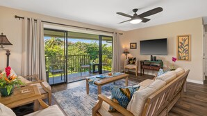 A spacious living room with a large sliding glass door leading to a balcony, a ceiling fan, a flat-screen TV, and a comfortable seating area. - Tropical Oasis - Stunning Views In Private Setting (Princeville)