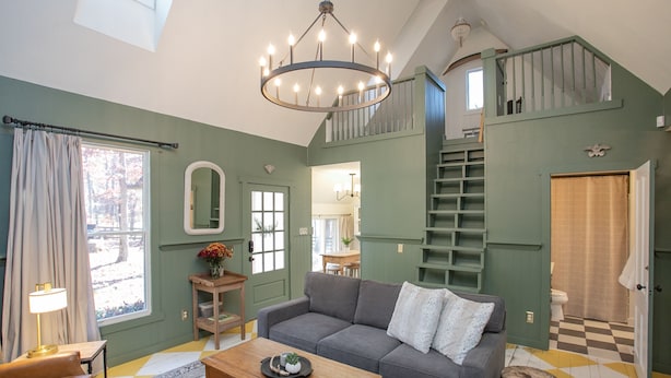 Ein geräumiges Wohnzimmer mit grauem Sofa, Holztisch und einer Kronleuchter. Eine Treppe führt zu einer oberen Ebene, und ein Fenster lässt Tageslicht herein.