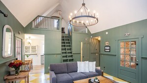Ein geräumiges Wohnzimmer mit grauem Sofa, Holztisch und einer Deckenleuchte. Eine Treppe führt zu einer oberen Ebene, und eine Tür mit Fenster ist sichtbar.