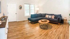 A living room with a dark blue sectional sofa, a round coffee table, wooden flooring, a white door, a window, and a floor lamp.