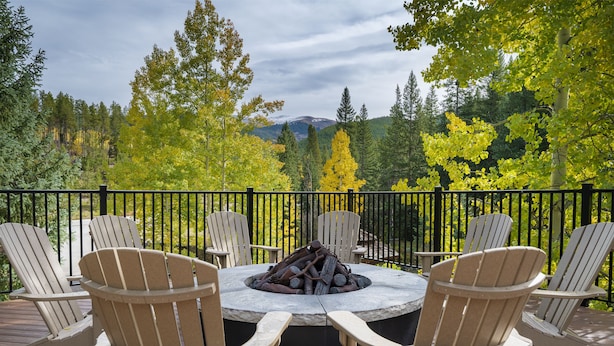 A deck with Adirondack chairs around a stone fire pit, overlooking a forested area with mountains in the distance.