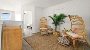 Un salon lumineux et moderne, avec des meubles en osier, une grande plante et une vue sur l’extérieur.
