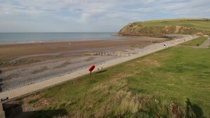 A coastal beach with a paved walkway, grassy areas, and a hill in the background.