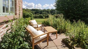 A patio with a wooden table and two cushioned chairs, a brick wall, and a view of a green field and trees.