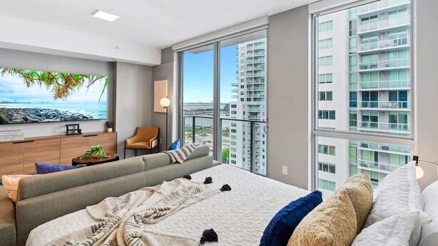 A modern bedroom with a large bed, a sofa, a TV, and a view of high-rise buildings. - Downtown Miami 1BR Condo with Stunning Ocean Views (Miami)