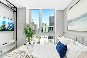 Quarto moderno com uma cama grande, uma televisão, uma planta e vista para a cidade por uma janela ampla.
