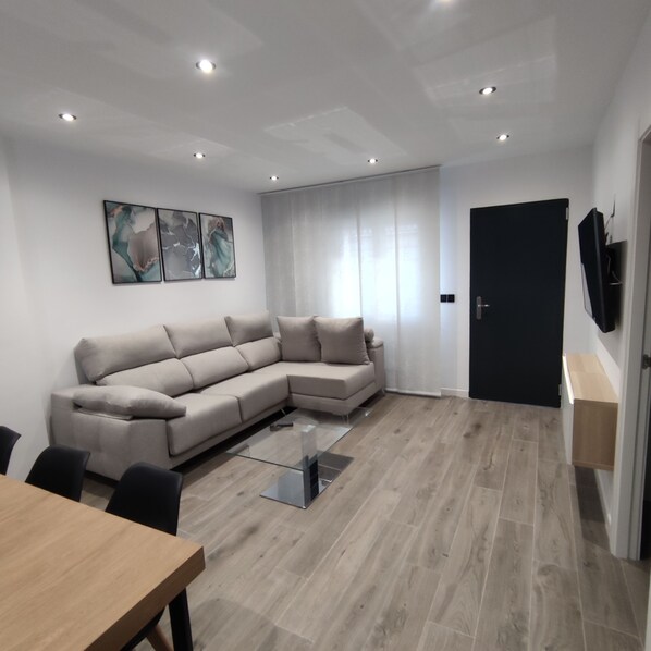 Un salon moderne avec un canapé d’angle beige, une table basse en verre, une table à manger en bois et une porte noire.
