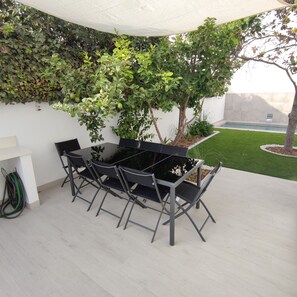 Une terrasse avec une table et des chaises, entourée de verdure et d’une petite pelouse.