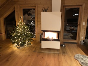 A cozy living room with a wooden fireplace, a Christmas tree, and a large window.