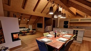 Un coin repas chaleureux et rustique, avec une table en bois dressée pour quatre personnes, une cheminée et une cuisine en arrière-plan.