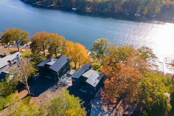 An aerial view of a lakeside property with modern buildings, a dock, and surrounding trees.