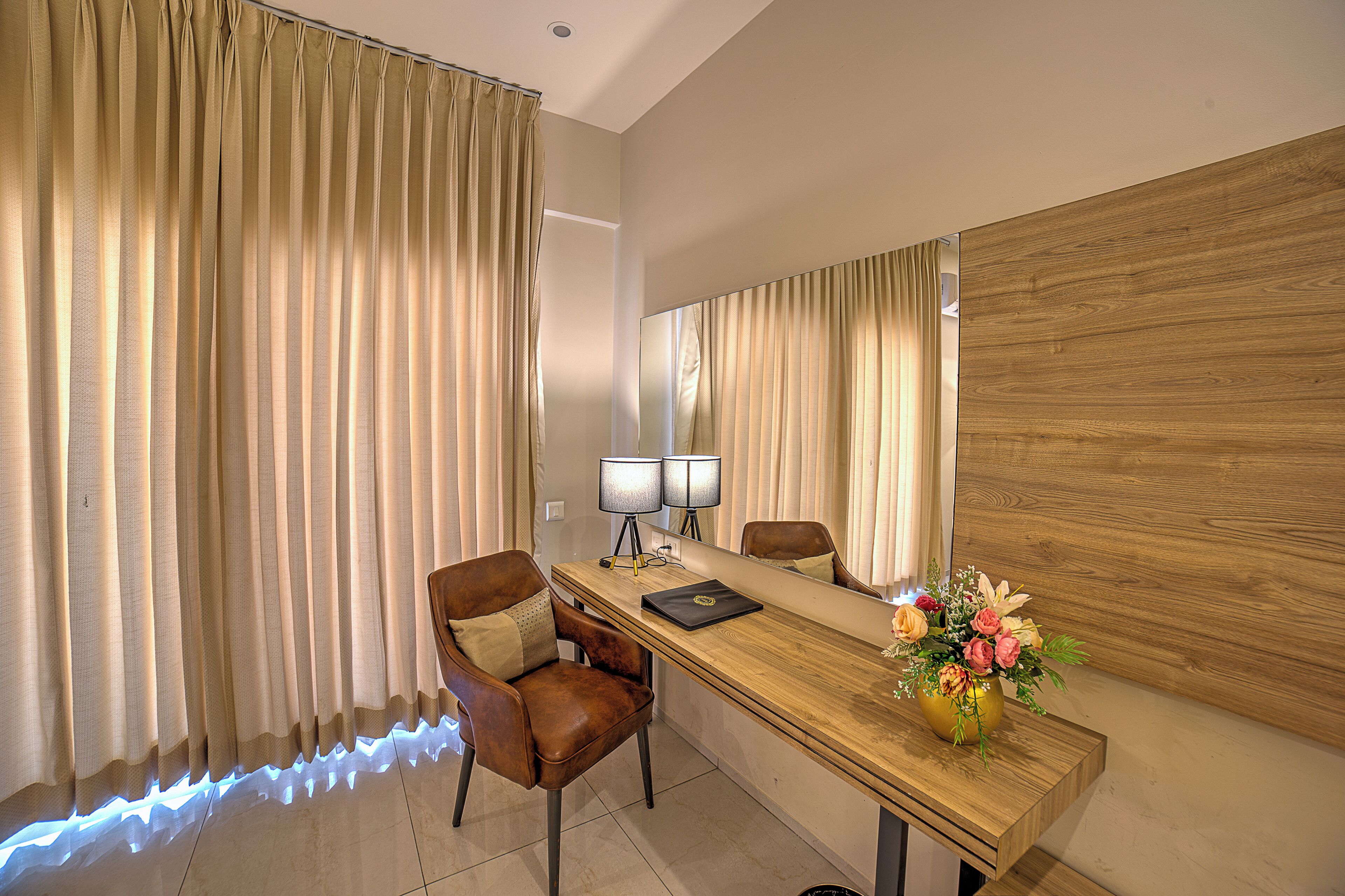 Quarto de hotel com escrivaninha de madeira, uma cadeira, um espelho e um vaso de flores.
