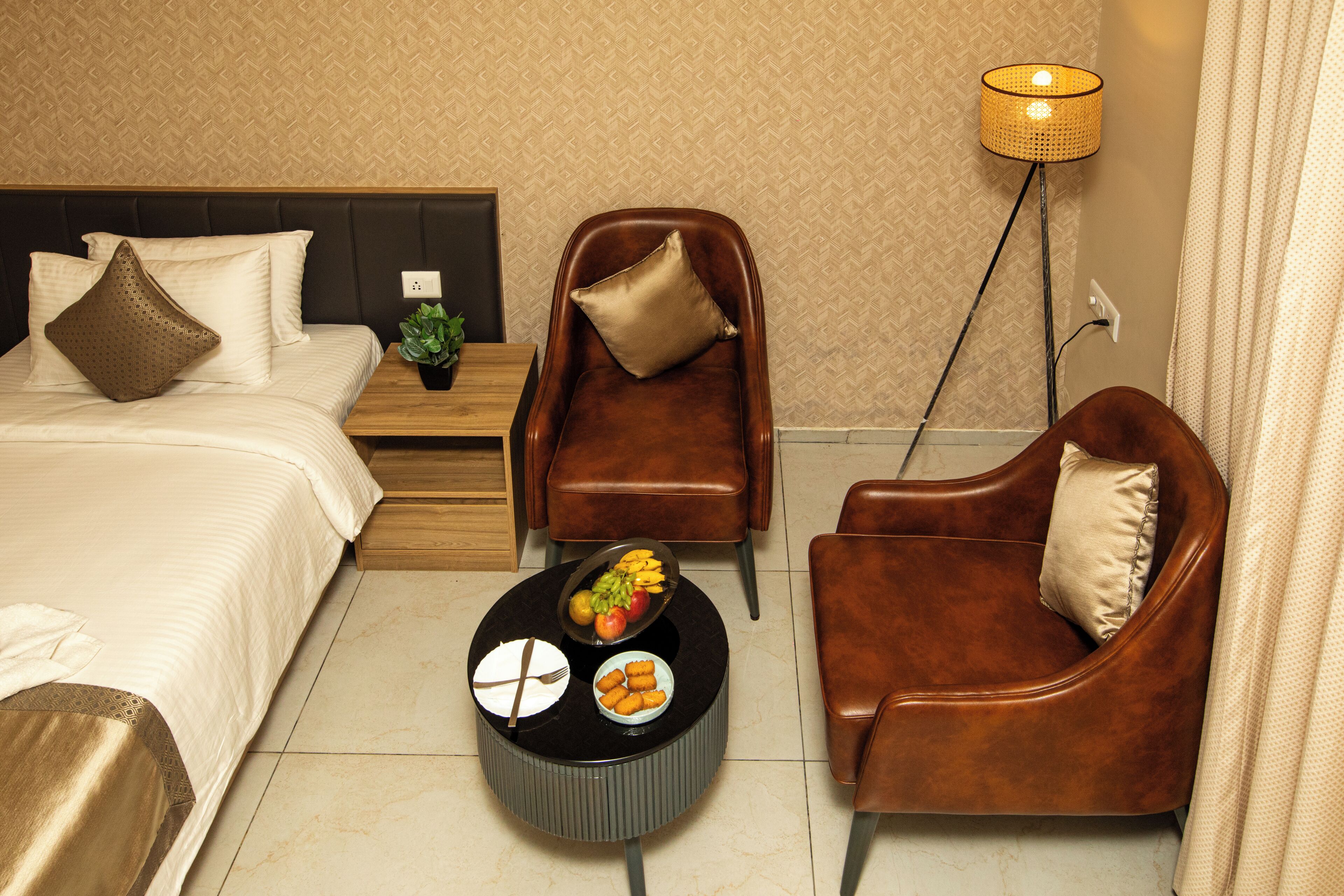 Habitación de hotel con una cama, dos sillones de cuero marrón, una mesita redonda con comida y una lámpara de pie.