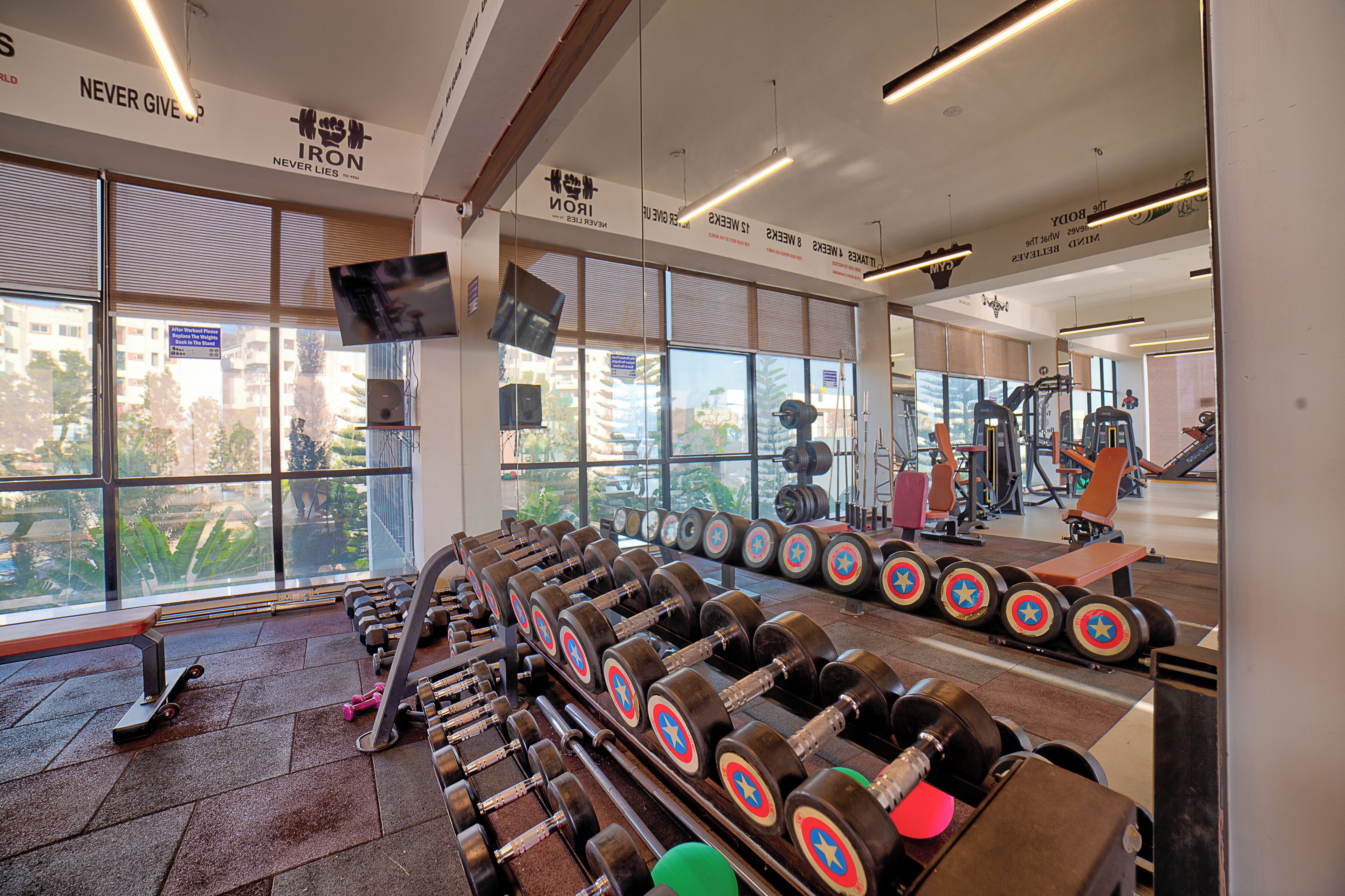A gym with rows of dumbbells, exercise equipment, and large windows offering a view of buildings and trees.