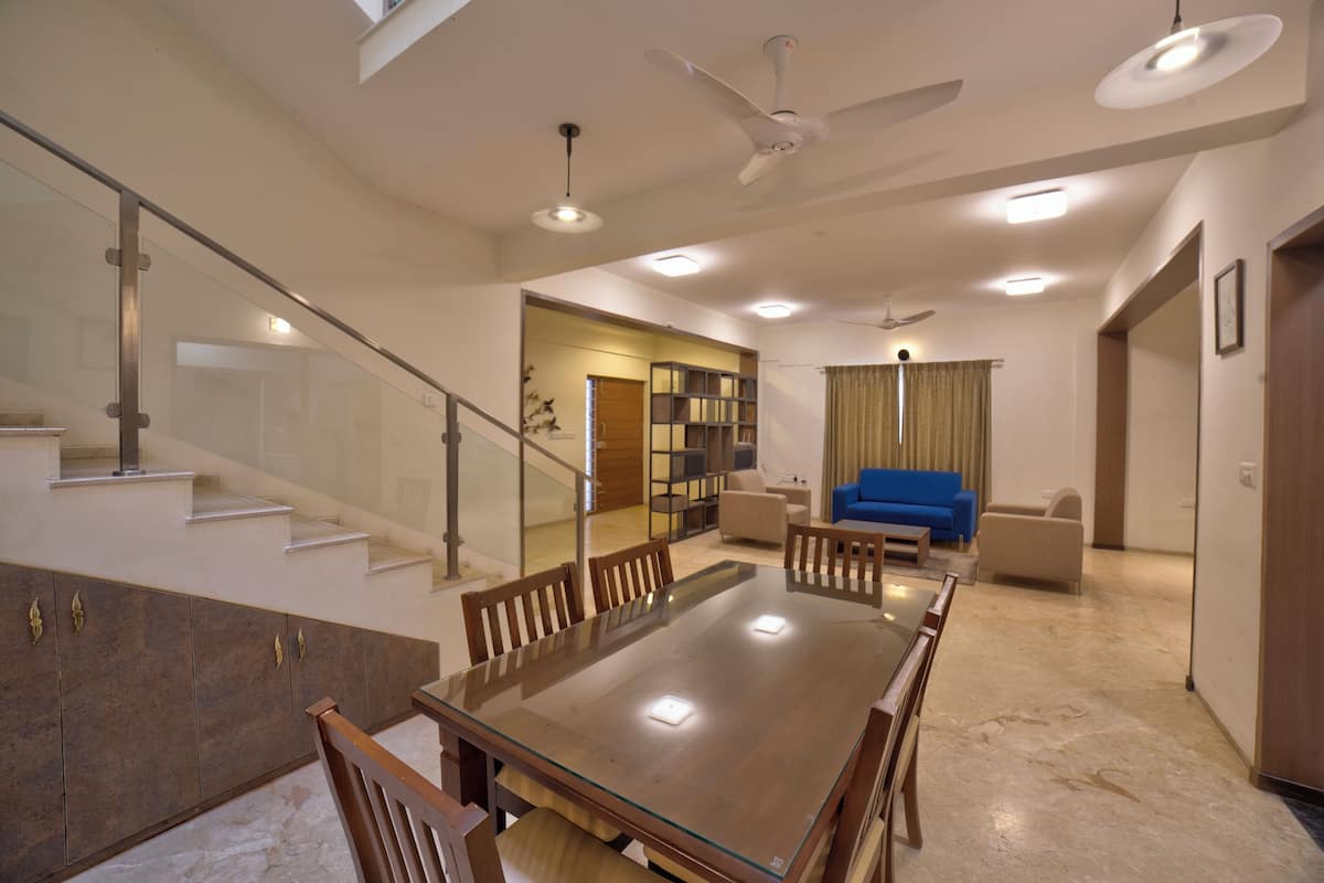 A spacious living area with a glass-top dining table surrounded by chairs, a blue sofa, and a staircase with glass railings.