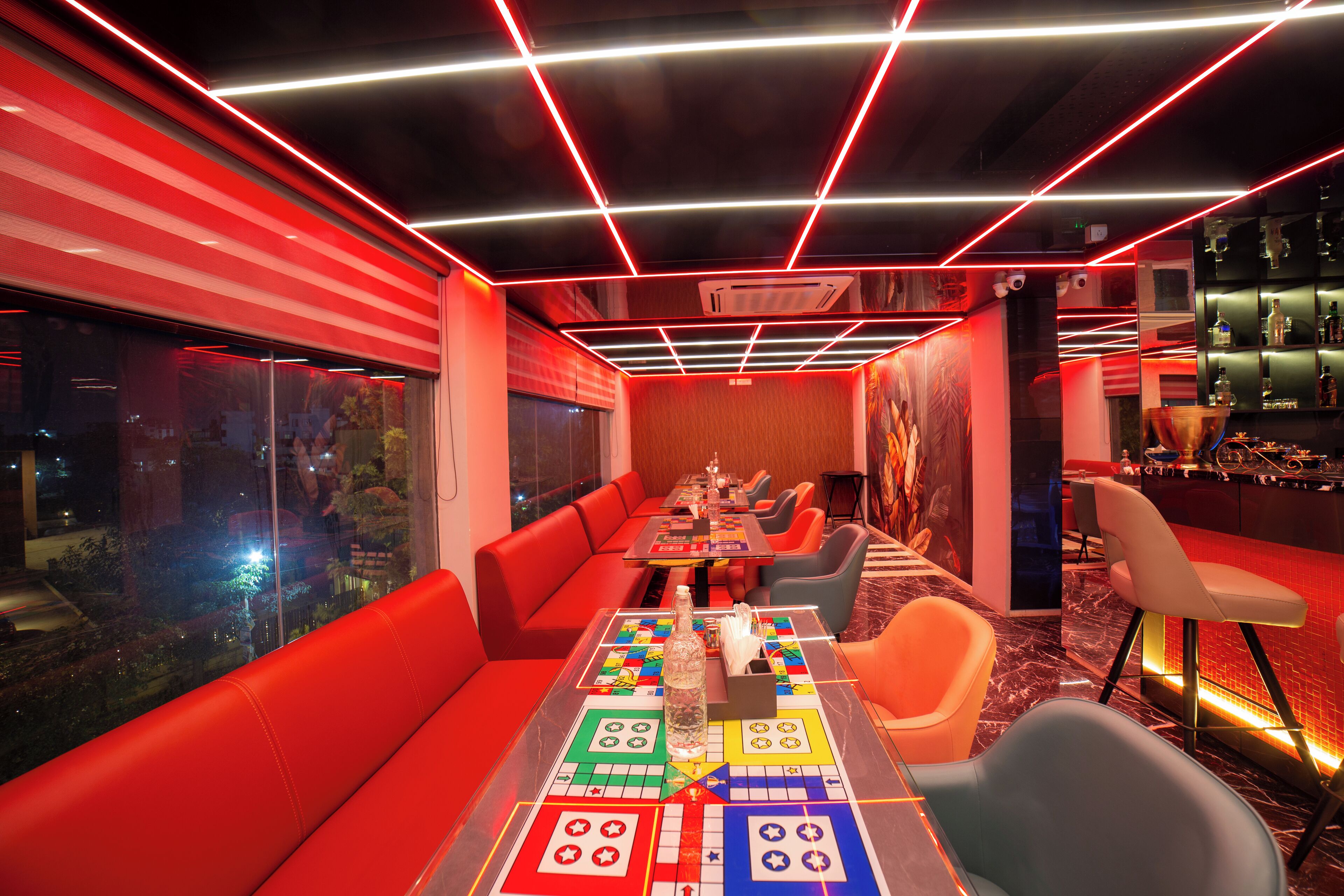 A modern bar area with red seating, a long table with a board game, and a bar counter with shelves of bottles.