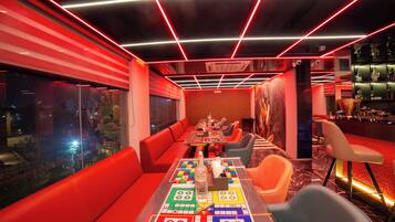 A modern bar area with red seating, a long table with a board game, and a bar counter with shelves of bottles.