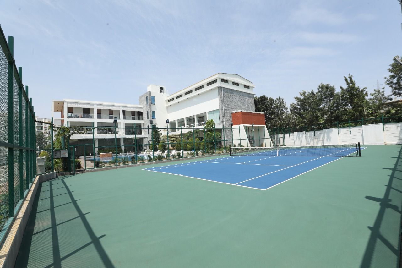 Una cancha de tenis con una red, rodeada por una cerca verde y un edificio de varios pisos con techo rojo.