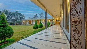 A covered walkway with a patterned railing, leading to a building with multiple doors and a well-maintained lawn with trimmed bushes. - VRR Hotel's And Convention Centre (Anekal)