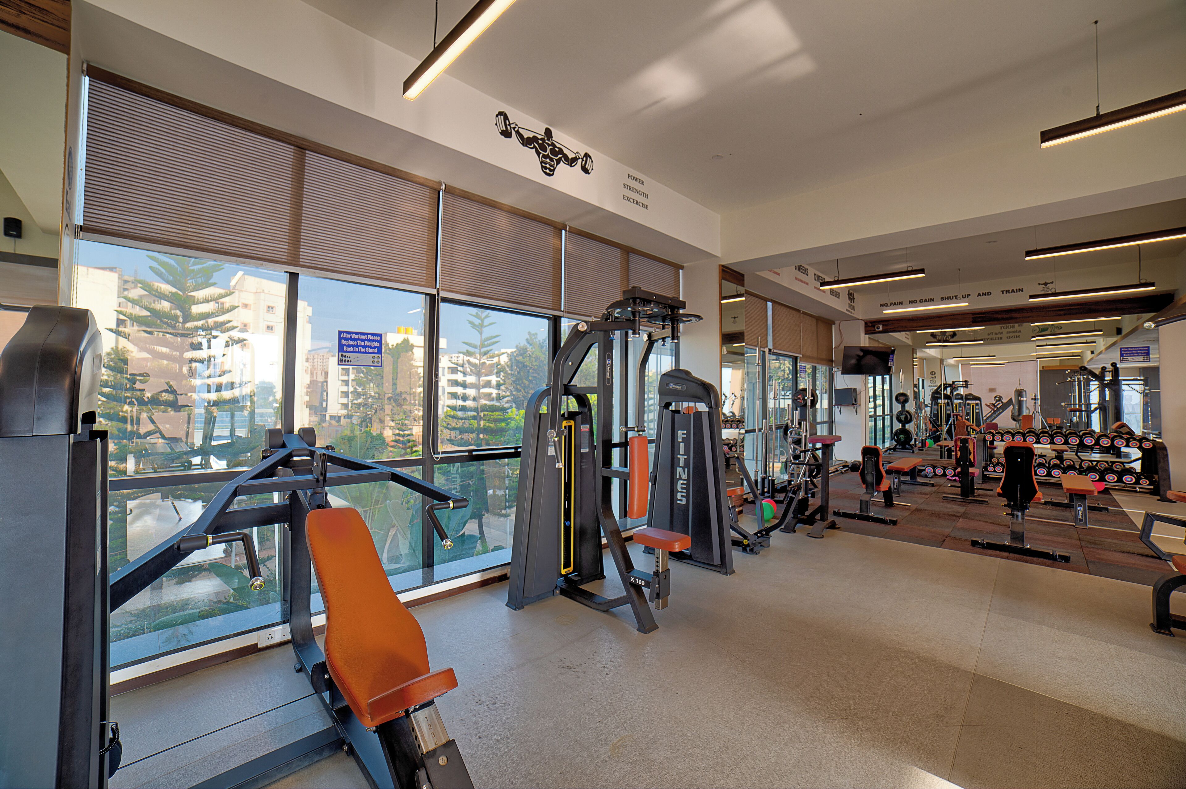 A modern gym with various exercise equipment, large windows offering views of buildings, and a clean, well-lit interior.