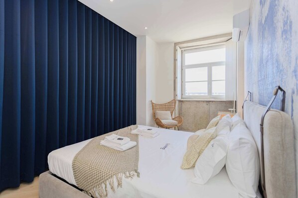 Une chambre d’hôtel moderne avec un grand lit, une fenêtre, un fauteuil en osier et un climatiseur fixé au mur.
