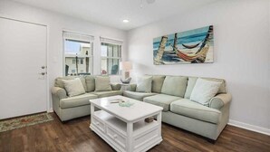 A living room with a sectional sofa, a white coffee table, and a hammock painting on the wall.
