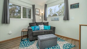 Un salon chaleureux avec un canapé gris, une petite table d’appoint et un pouf gris. Il y a deux fenêtres avec des rideaux, une lampe fixée au mur et un tableau encadré.
