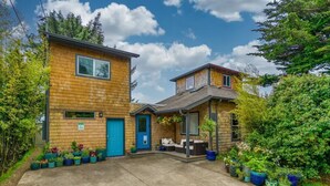 Une maison en bois à deux étages, avec une porte bleue et une véranda couverte aménagée avec du mobilier d’extérieur.