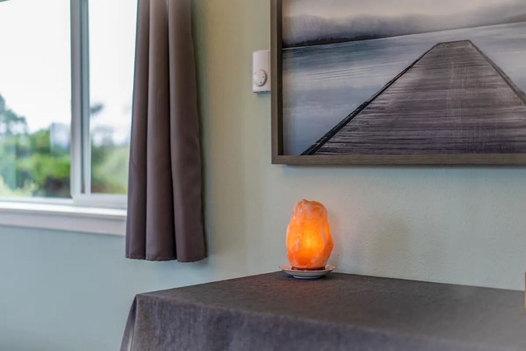 A room with a salt lamp on a table, a painting on the wall, and a window with curtains.
