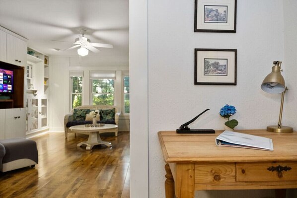 Una habitación con piso de madera, ventilador de techo, sofá blanco, mesa de centro y escritorio con lámpara y planta.