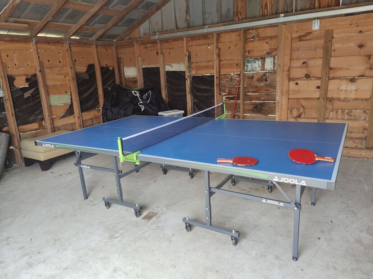 A ping pong table with two paddles, a table tennis ball, and a table tennis paddle in a wooden room with a metal roof.