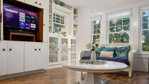 Una habitación con una televisión mostrando servicios de transmisión, un armario blanco con puertas de cristal, un sofá de mimbre con cojines estampados y una mesa redonda con un jarrón.