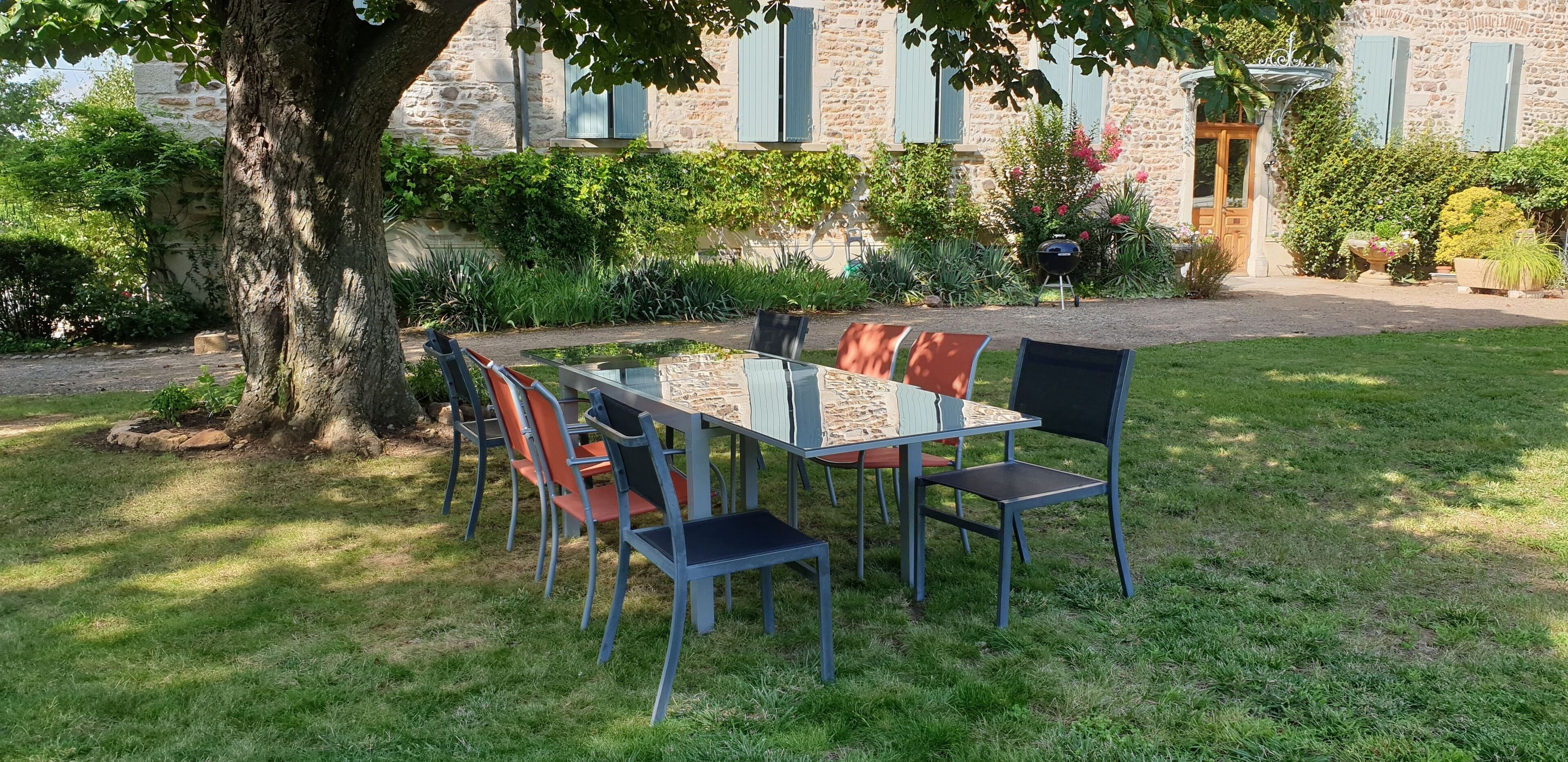 Een eethoek met een tafel en stoelen buiten, omringd door een tuin en een stenen gebouw met luiken.