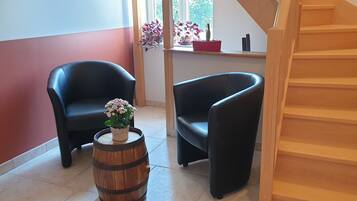 Una stanza con due poltrone nere, una botte di legno e un tavolino con una pianta in vaso. Si intravede una scala in legno a destra.