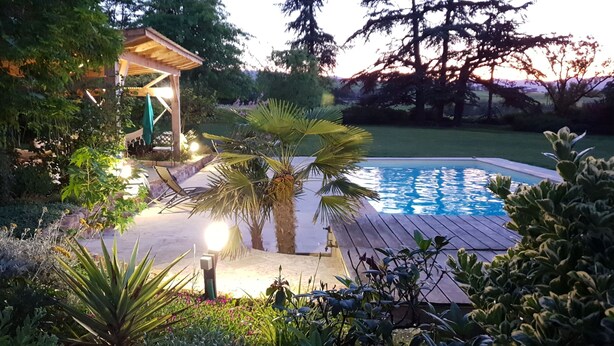 Una piscina con terraza de madera, rodeada de exuberante vegetación y un pabellón.
