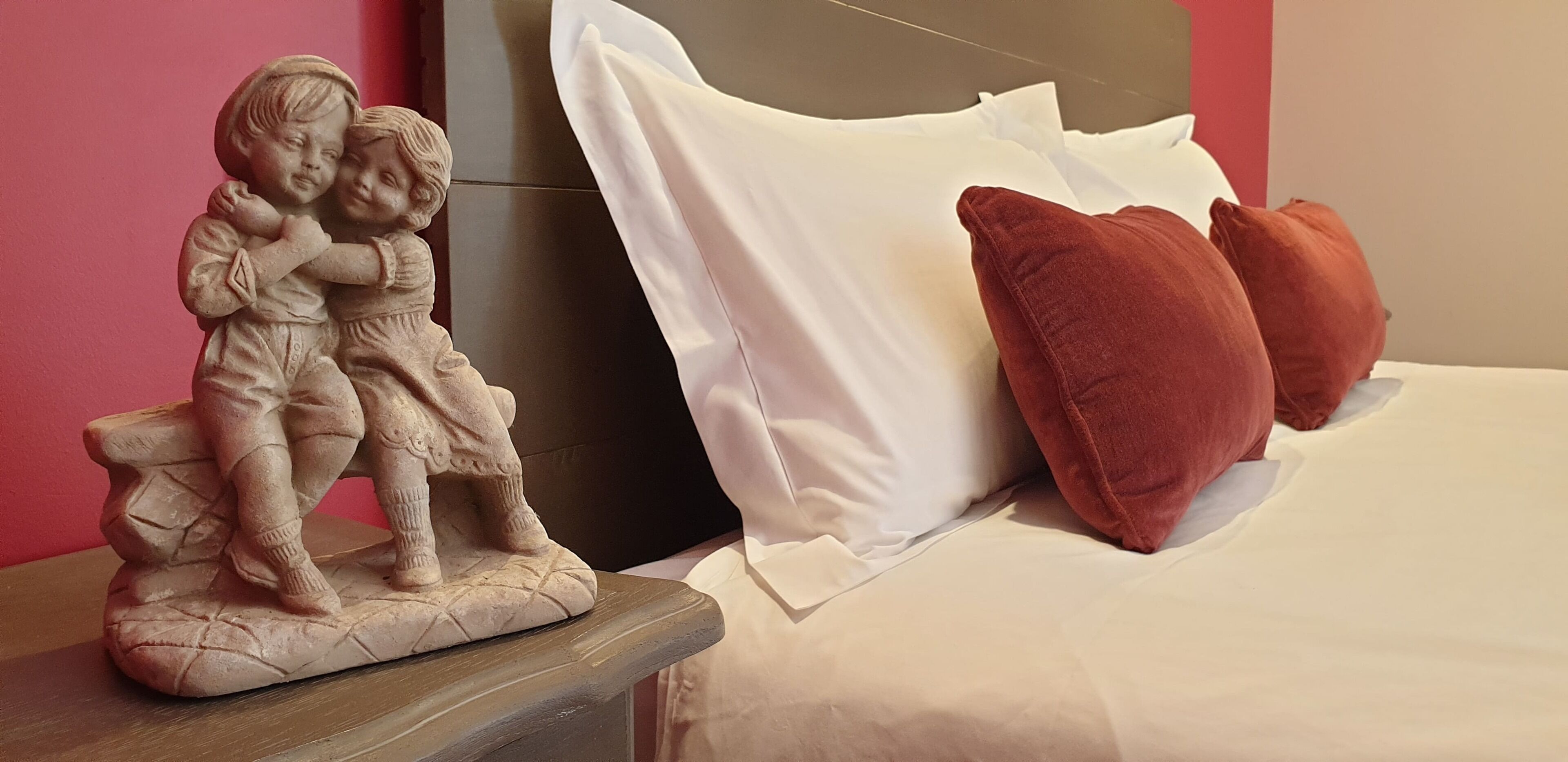 Un lit avec des oreillers blancs et bruns, une table de chevet ornée d’une sculpture représentant deux enfants, et un mur rouge.