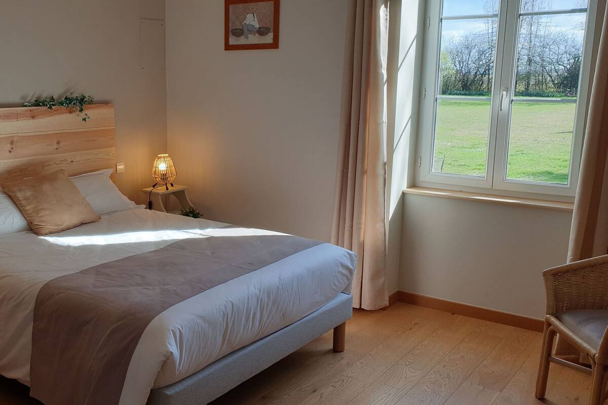 Un dormitorio con cabecera de madera, cama con ropa de cama blanca y beige, dos lámparas de noche, una silla y una ventana con vista a un área con césped.