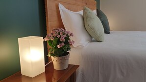 Una cama bien tendida con ropa blanca, un cabecero de madera y una mesita de noche con una lámpara blanca y una planta en maceta con flores rosadas.