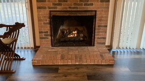 Une cheminée en briques avec un feu, un fauteuil en bois et une vue par la fenêtre.