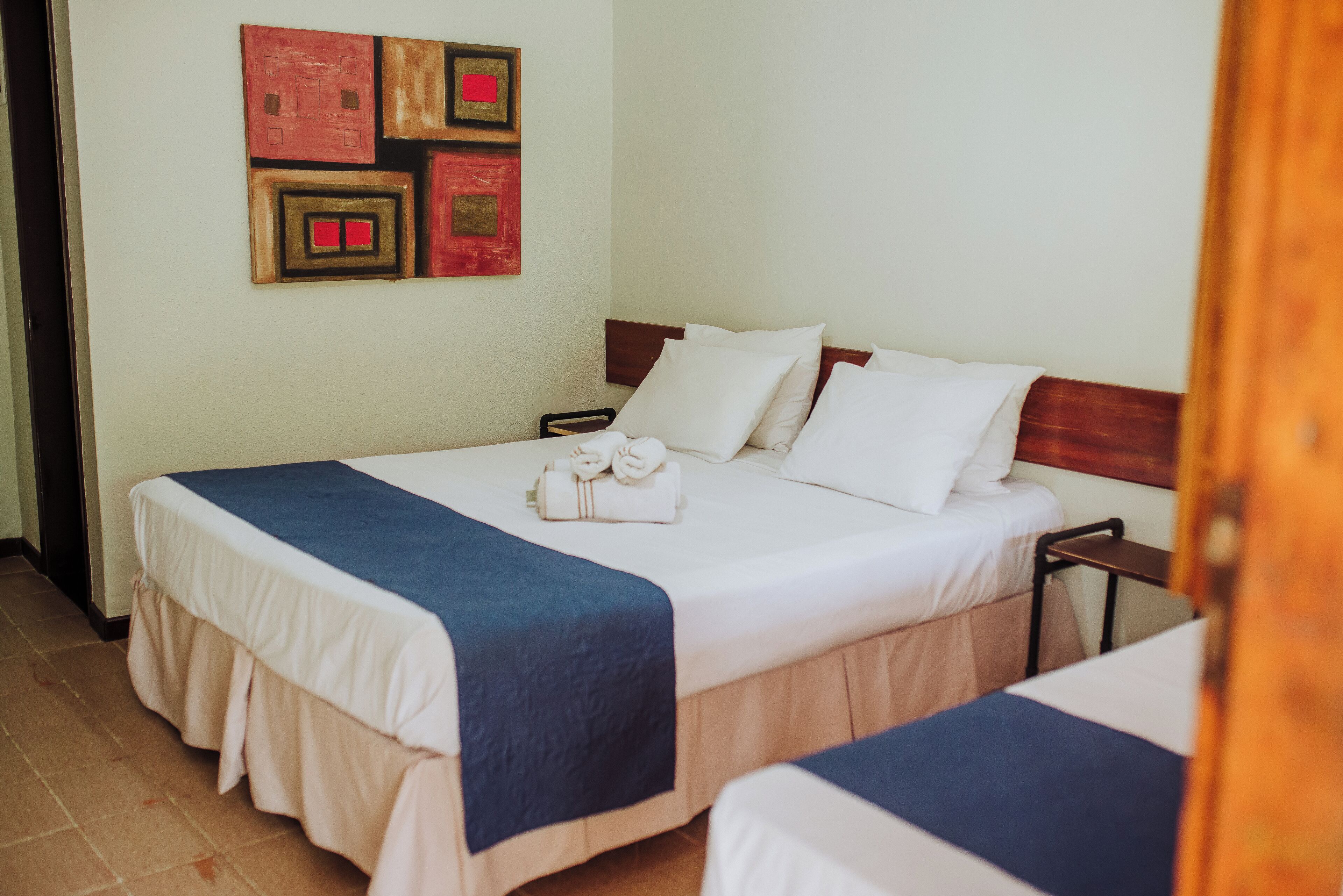 A hotel room with two beds, a wooden headboard, white bedding, a blue accent on the bedspread, and a wall art piece.