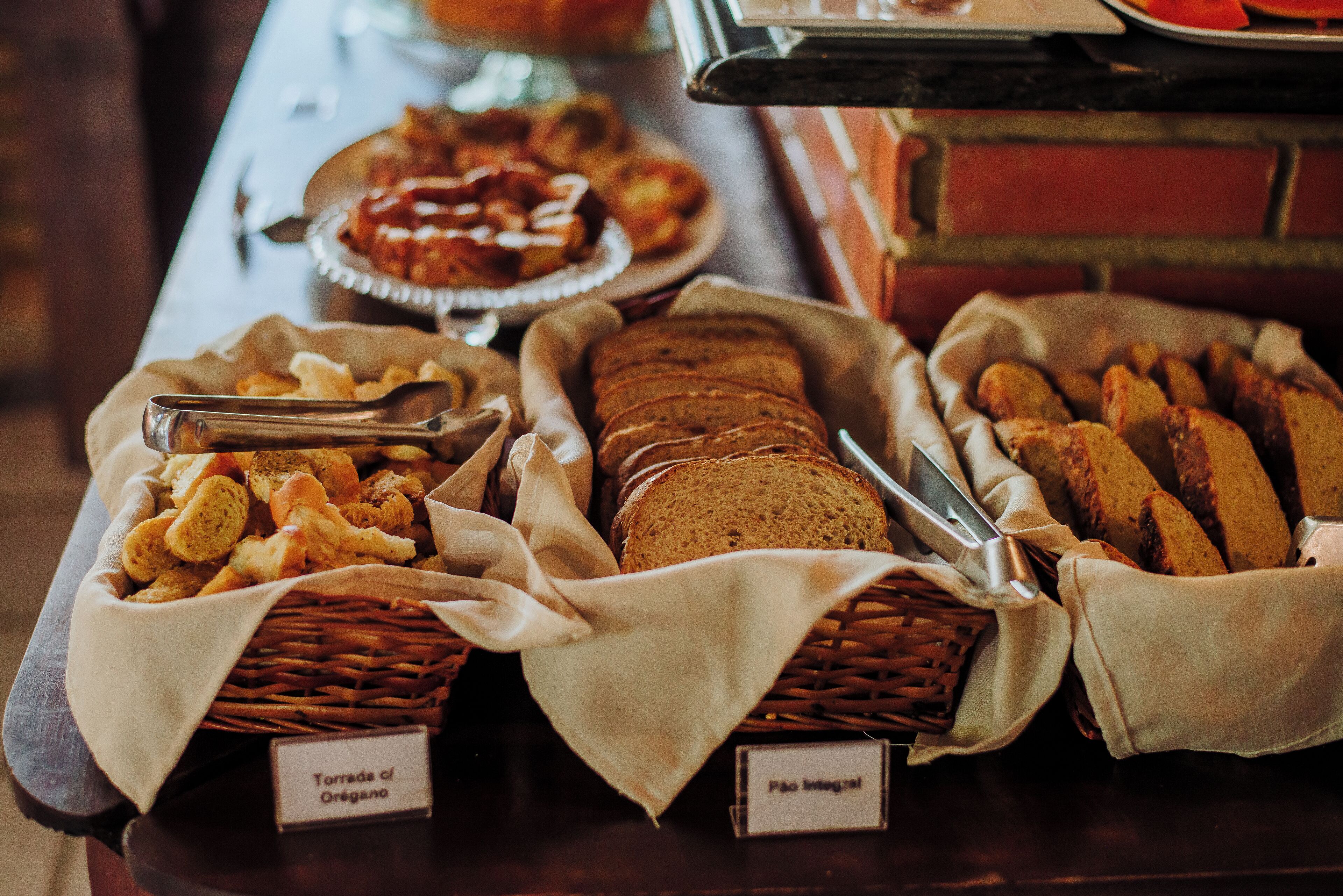 A buffet table with labeled baskets of Tortada de Oregano and Pão Integral.