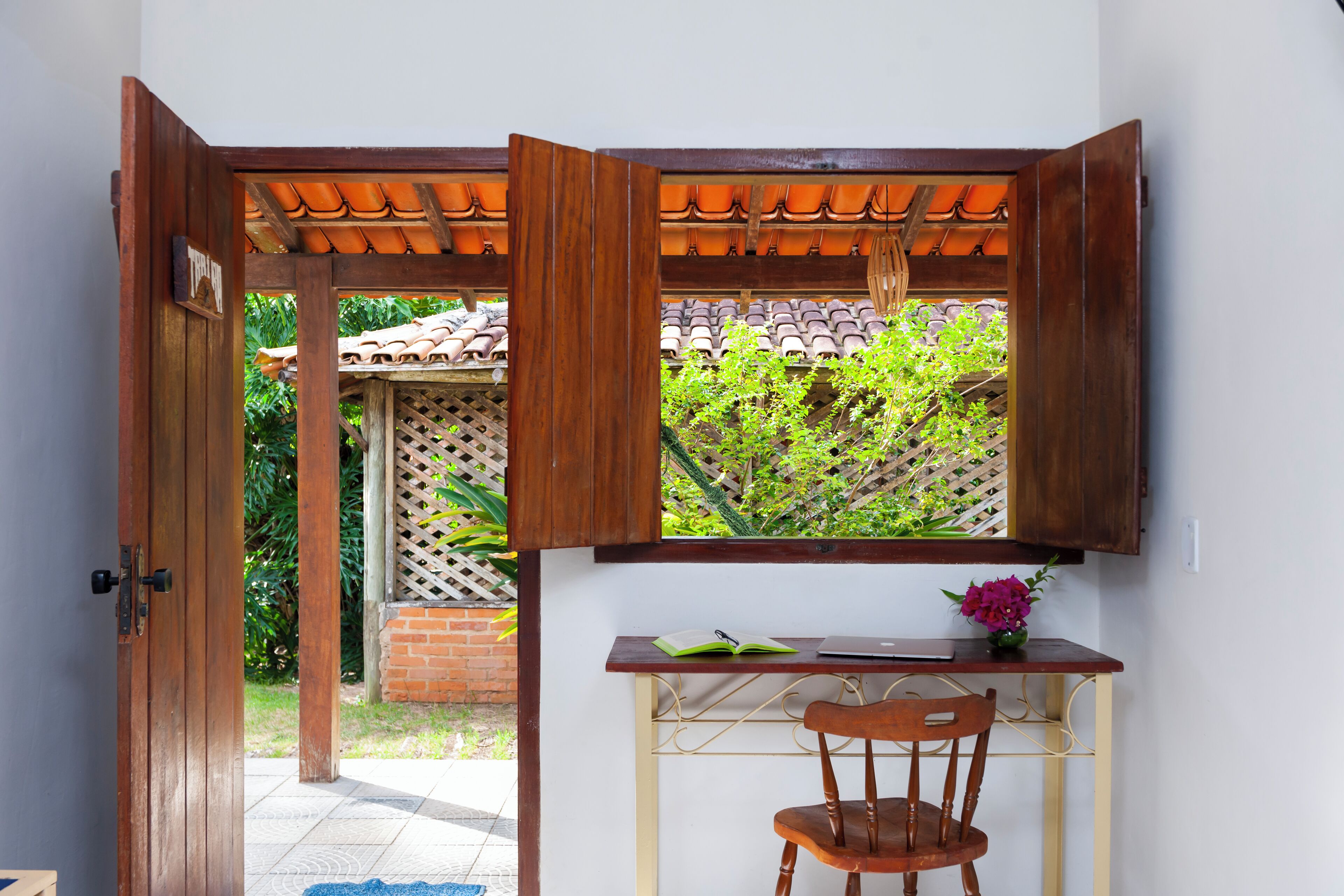 A room with a wooden desk, a chair, a window with a view of greenery, and a door leading outside.