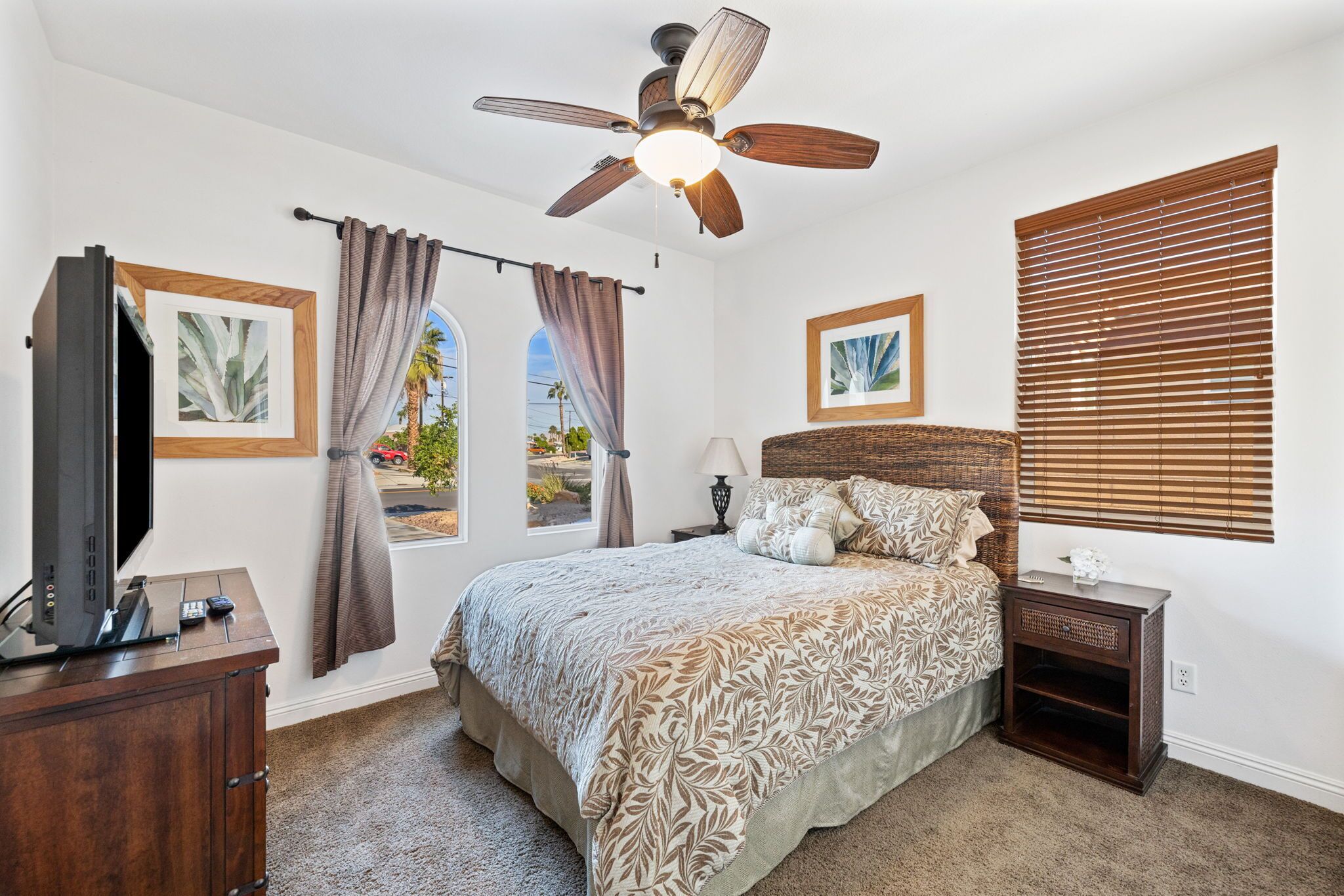 A bedroom with a bed, a ceiling fan, a TV on a wooden cabinet, a nightstand, and a window with blinds.